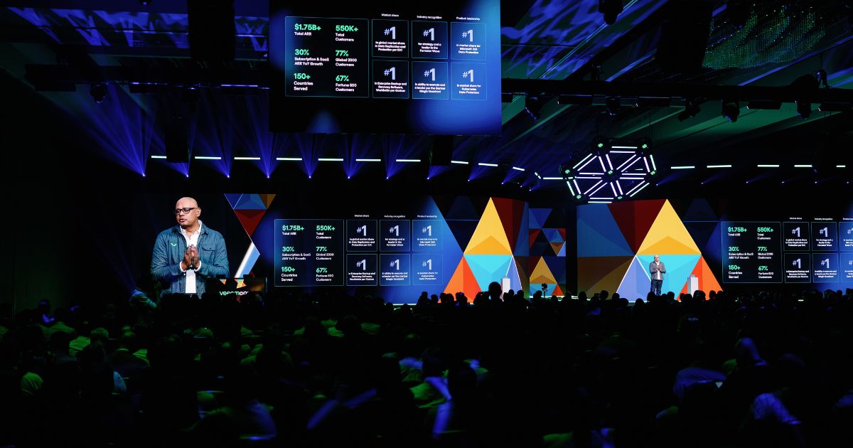 Uma conferência de tecnologia ocorre num auditório amplo e escuro, iluminado por luzes LED azuis e verdes. Um orador careca, com óculos e casaco azul-escuro, está em pé no palco a apresentar dados projetados em grandes ecrãs digitais. O fundo do palco é decorado com painéis coloridos em formas geométricas. Nos ecrãs lê-se informação sobre crescimento financeiro, estatísticas de clientes, presença global e liderança de mercado, com destaque para números como "$1.75B+ Total ARR", "550K+ Clientes" e "150+ Países". A audiência aparece em silhueta, atenta à apresentação.