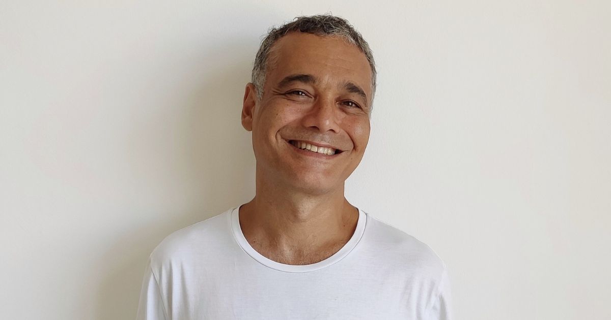 Sidarta Ribeiro sorri levemente em frente a uma parede clara. Ele veste uma camiseta branca e tem cabelo curto e grisalho. Foto: Luiza Mugnol Ugarte (tecnologia)