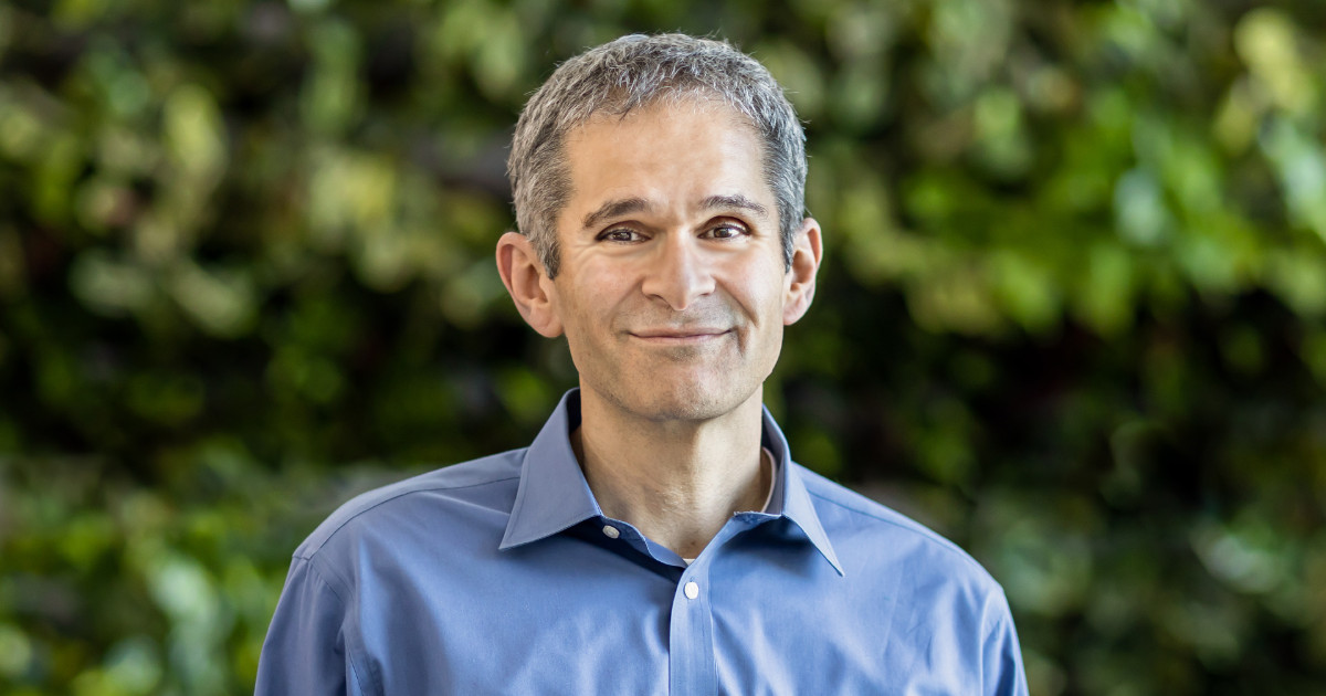 Amin Vahdat, homem de meia-idade com cabelos grisalhos curtos, vestindo uma camisa azul e sorrindo levemente, posando em frente a um fundo desfocado com folhagem verde. Ele é Vice-presidente de Aprendizado de Máquina, Sistemas e Inteligência Artificial em Nuvem do GOogle Cloud (agentes)