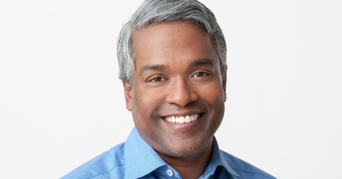Homem sorridente, de pele morena, cabelo curto e grisalho, vestindo camisa azul. Esta é uma foto de Thomas Kurian, CEO do Google Cloud, em frente a um fundo branco.