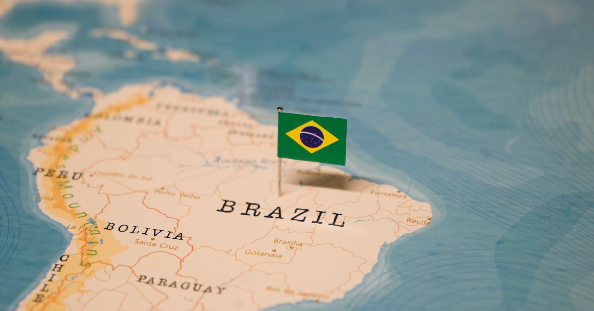 Mapa da América do Sul com foco no território brasileiro, onde uma pequena bandeira do Brasil está fincada sobre a área central do país, destacando visualmente sua localização geográfica (Inovação)