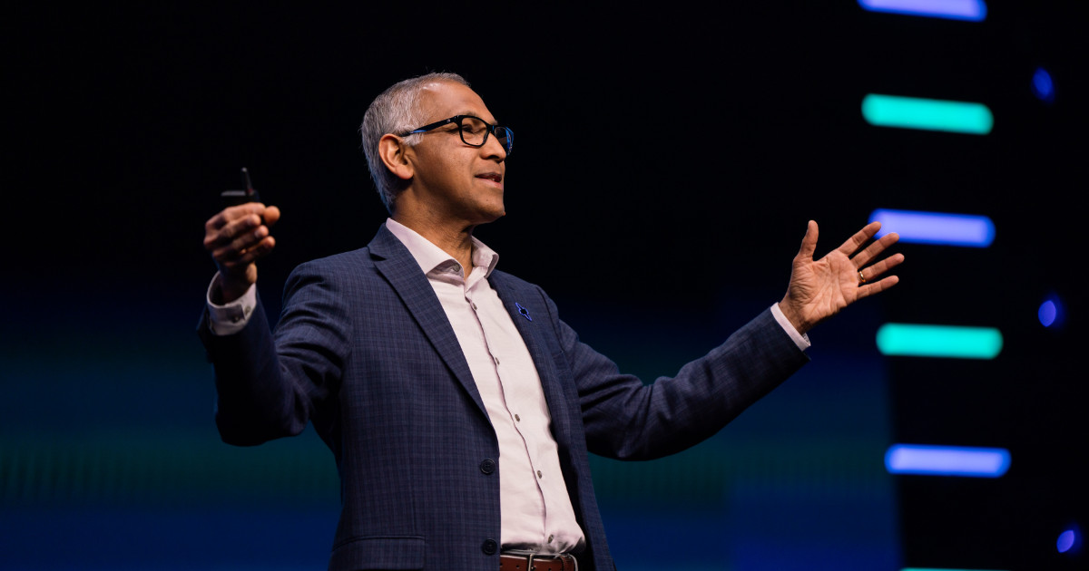Rajiv Ramaswami, CEO da Nutanix, aparece em um palco durante uma apresentação. Ele está vestindo terno azul xadrez, camisa social branca e óculos, com os braços abertos em gesto expressivo, segurando um controle remoto em uma das mãos. Ao fundo, o cenário é escuro com luzes de LED coloridas em listras horizontais.