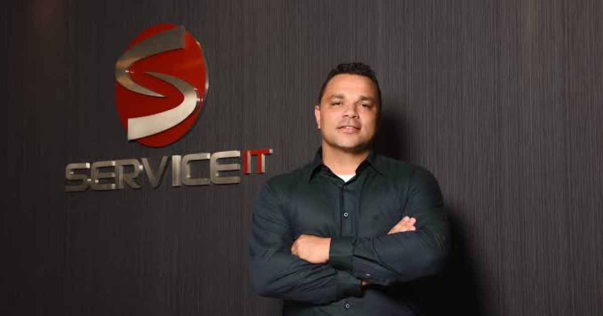 Homem de braços cruzados em frente a uma parede escura com o logótipo metálico da empresa “SERVICE IT”. O logótipo inclui um símbolo vermelho e prateado em forma de “S” estilizado. O homem está vestido com uma camisa escura e tem uma expressão confiante e profissional.