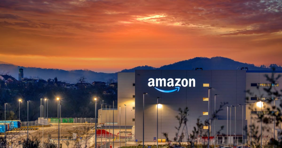 Centro logístico da Amazon ao entardecer, com o logótipo da empresa iluminado na fachada de um grande edifício industrial cinzento. O céu exibe tons vibrantes de laranja e vermelho ao pôr do sol, com colinas escuras e silhuetas de árvores ao fundo. À frente do armazém, há postes de iluminação acesos e contentores coloridos, sugerindo uma zona de carga e descarga. O ambiente mistura a grandiosidade do cenário natural com a modernidade das instalações da empresa.