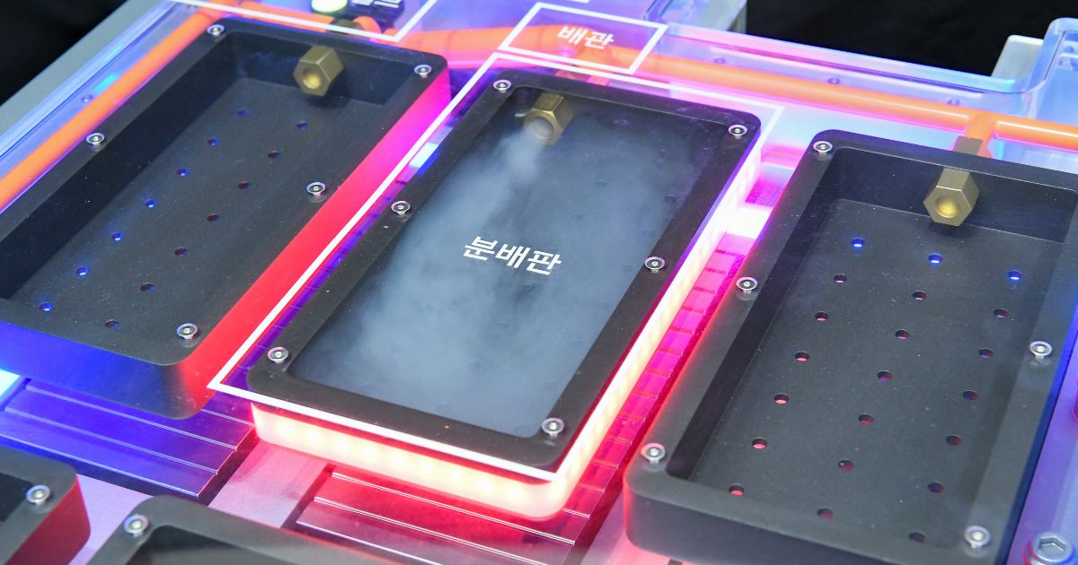 Uma estrutura retangular tecnológica com iluminação LED nas bordas, exibindo três compartimentos pretos com furos circulares em suas superfícies. O compartimento central emite uma névoa ou vapor branco e possui texto em coreano ("분배판", que significa "placa de distribuição"). Os outros dois compartimentos estão apagados, mas compartilham o mesmo design. Há um tubo ou bico metálico na parte superior de cada compartimento, sugerindo entrada ou saída de ar ou líquido. A estrutura parece fazer parte de um sistema de demonstração ou equipamento industrial de alta tecnologia (bateria)