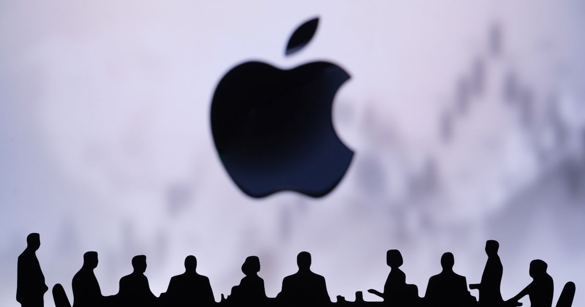 Silhuetas de pessoas sentadas à volta de uma mesa de reunião, em frente ao logótipo da Apple em destaque ao fundo, com um gráfico desfocado ao fundo sugerindo contexto financeiro ou empresarial.