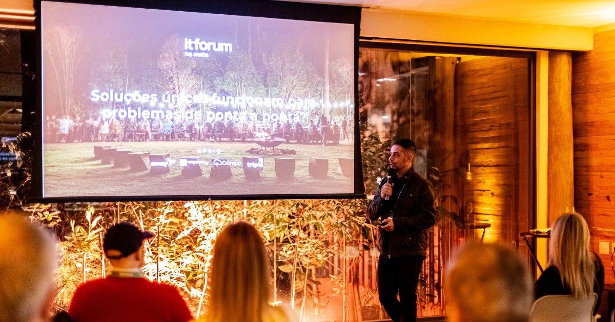 Homem em pé com microfone na mão a fazer uma apresentação noturna durante um evento do itforum, com pessoas sentadas a assistir e um ecrã grande ao fundo com a frase 'Soluções únicas de fundo para resolver problemas de ponta a ponta', tecnologia