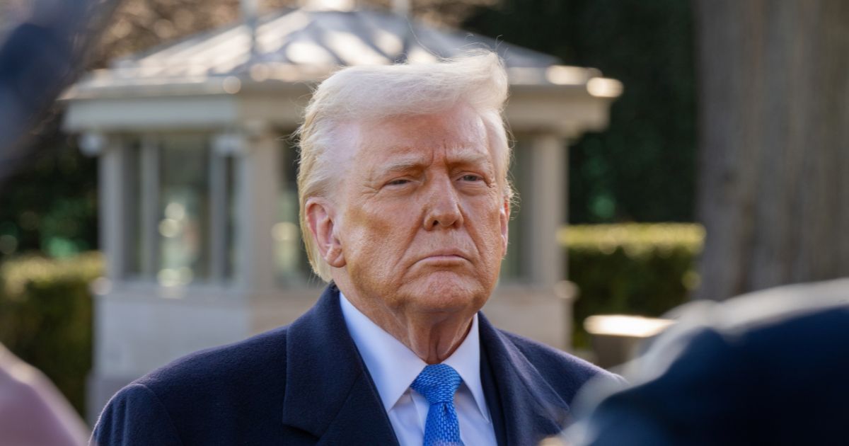 Um homem de expressão séria, com cabelo loiro penteado para trás e pele com tonalidade bronzeada, veste um sobretudo escuro, camisa branca e gravata azul. Ele está ao ar livre, em frente a uma estrutura clara com janelas, possivelmente parte de um edifício governamental ou um jardim formal. O fundo é composto por vegetação e arquitetura clássica, sugerindo um ambiente institucional (donald trump, eua)