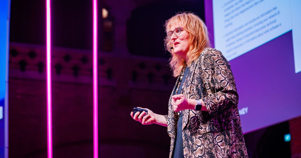 Mulher de cabelo loiro-ruivo, com óculos e vestida com um casaco de padrão de cobra, fala num palco iluminado com luzes cor-de-rosa. Ela segura um controlo remoto e usa um microfone auricular. Ao fundo, vê-se um ecrã de projeção com texto e o ambiente de auditório escuro, sugerindo um evento profissional ou tecnológico (futuro)