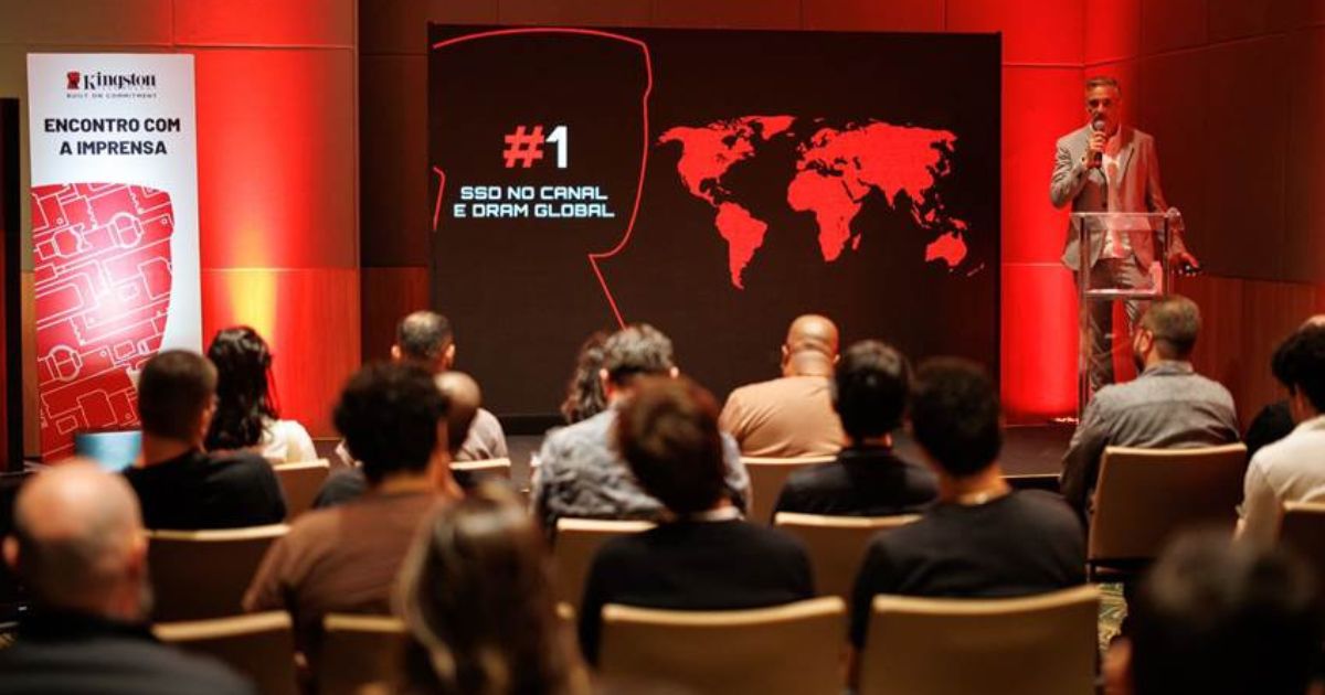 Fotografia de um evento corporativo da Kingston, com o título “Encontro com a Imprensa” num painel à esquerda. A imagem mostra uma plateia sentada assistindo a uma apresentação. À direita, um homem em fato cinzento está em pé, a falar ao microfone num púlpito transparente. Atrás dele, um grande ecrã preto e vermelho exibe um mapa-múndi e a frase “#1 SSD no canal e DRAM global”. O ambiente está iluminado com tons quentes, principalmente vermelhos, conferindo um tom sofisticado e moderno ao evento.