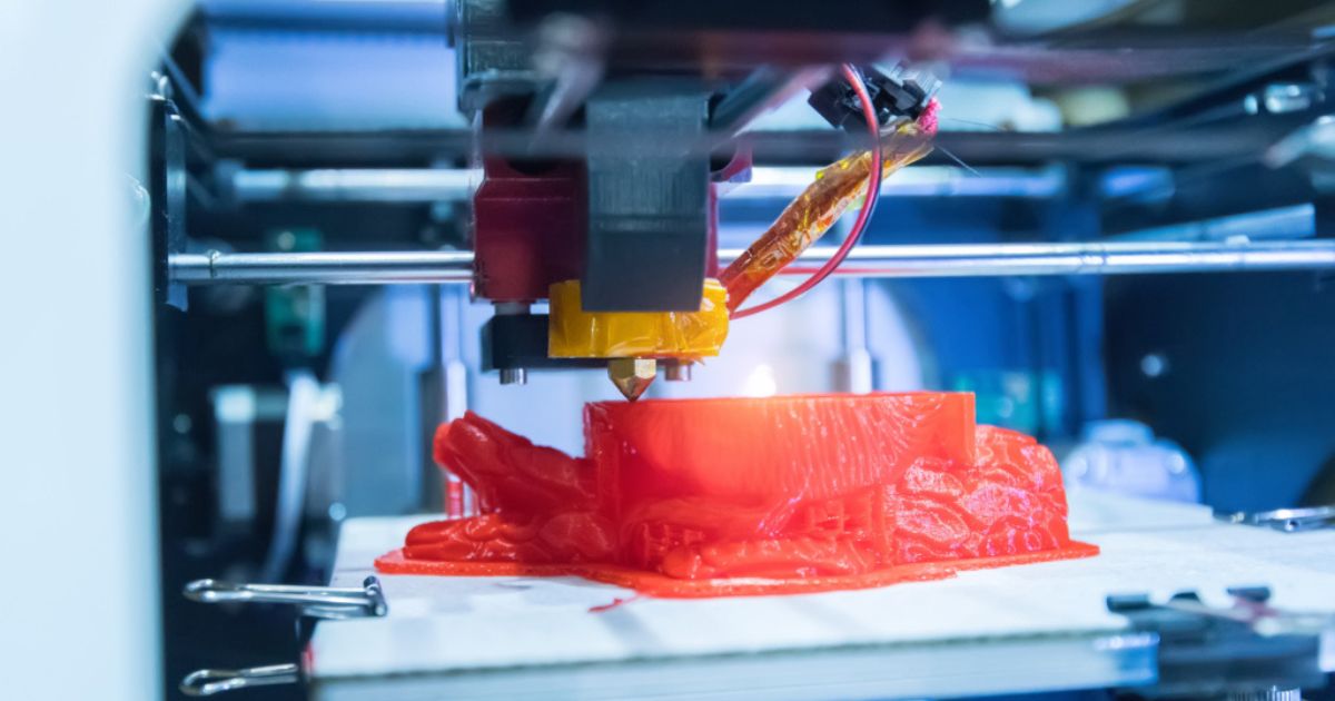 Close-up de uma impressora 3D em funcionamento, a criar um pulmão vermelho em plástico. A cabeça de extrusão está ativa, libertando o material em camadas sobre a plataforma de impressão. A imagem transmite inovação e tecnologia de fabrico digital.