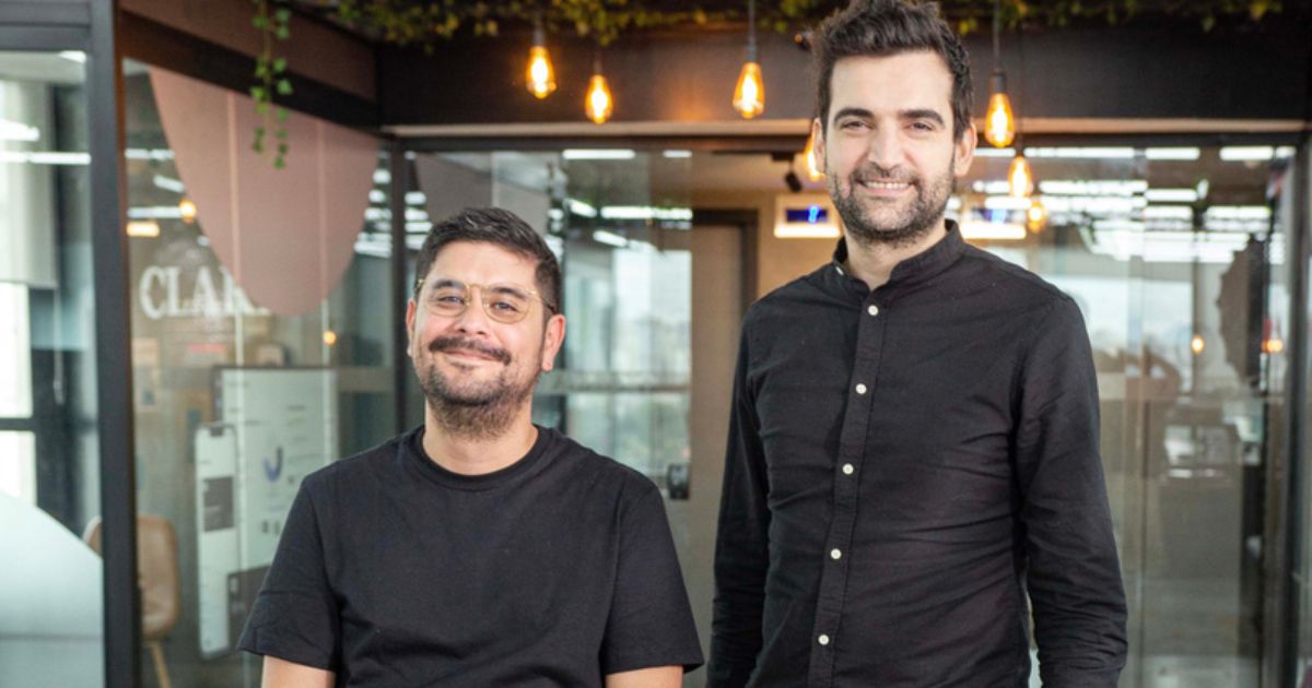 Dois homens a sorrir posam num ambiente de escritório moderno e iluminado. O homem à esquerda está sentado, usa óculos e uma t-shirt preta. O homem à direita está de pé, veste uma camisa preta de manga comprida. Ao fundo vê-se uma parede de vidro com iluminação suspensa e detalhes de plantas decorativas no teto.