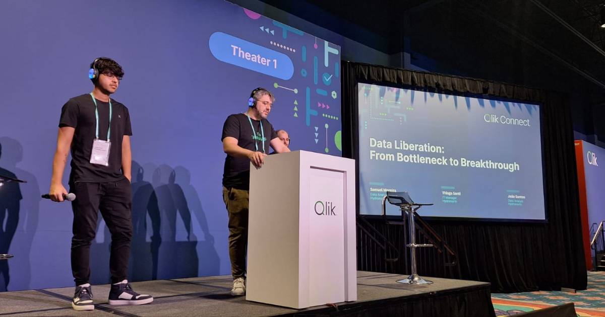Thiago Santi, gerente de TI da Hydronorth, no púlpito durante apresentação no evento Qlik Connect 2025. Ele está acompanhado de outros dois palestrantes no palco identificado como "Theater 1", com fones de ouvido e crachás. Ao fundo, o telão exibe o tema da apresentação: “Data Liberation: From Bottleneck to Breakthrough”.