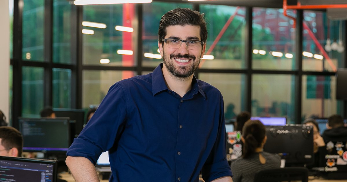 Thoran Rodrigues, CEO da BigDataCorp, está sorrindo e posando em um ambiente de escritório moderno, com várias pessoas ao fundo trabalhando em computadores. Ele veste uma camisa azul escura e óculos, e o ambiente tem amplas janelas de vidro com estrutura metálica visível (empresas)