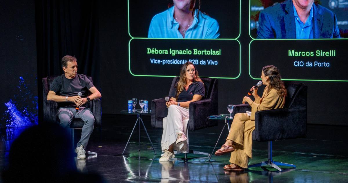 Marcos Sirelli, diretor de TI da Porto, e Débora Ignacio Bortolasi, vice-presidente B2B da Vivo, participam de um painel no IT Forum Trancoso 2025. Eles estão sentados em poltronas no palco, segurando microfones, enquanto conversam com uma mediadora. Ao fundo, há uma tela com os nomes e cargos dos participantes.