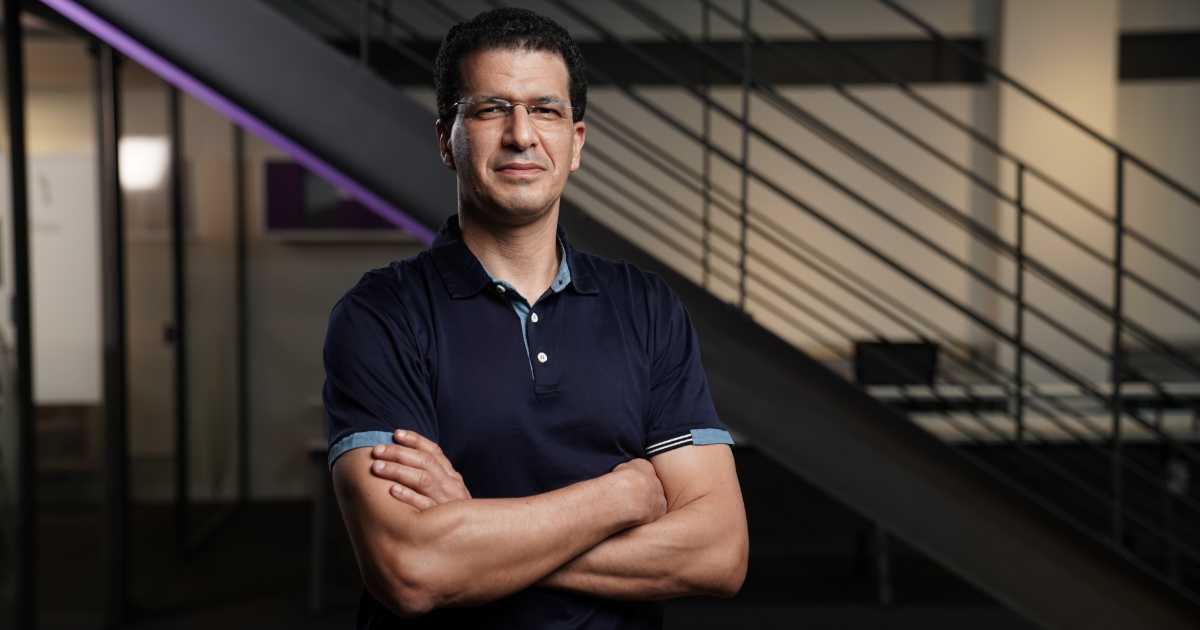 Homem de pele clara, cabelos curtos e escuros, usando óculos e camisa polo azul escuro, está de braços cruzados e olhando para a câmera com expressão confiante. Ele está em um ambiente corporativo moderno, com escada metálica e paredes de vidro ao fundo. A imagem mostra Youseef Lahrech, presidente e COO do Nubank