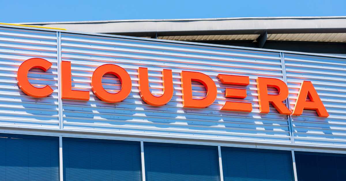 Fachada de prédio com o logotipo da empresa Cloudera em letras grandes e laranja, fixado em uma estrutura metálica prateada. A imagem foi tirada durante o dia, com céu azul ao fundo e janelas espelhadas na parte inferior da fachada