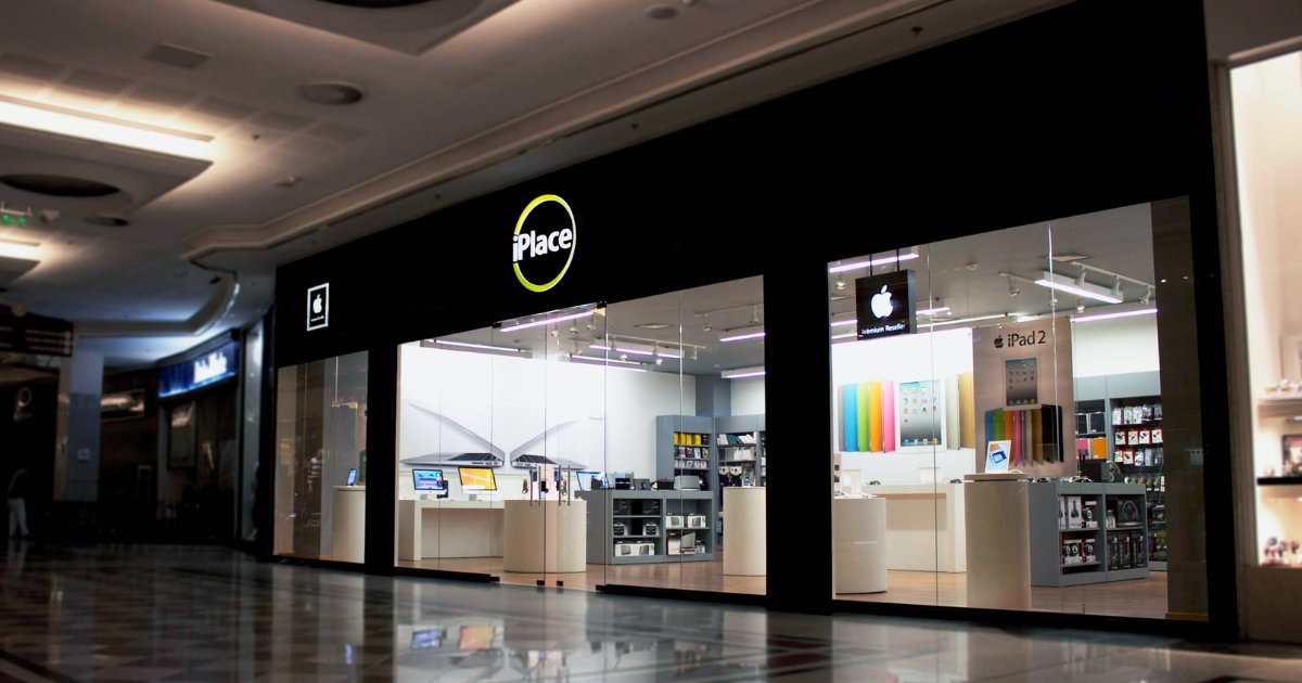 Fachada de uma loja iPlace em um shopping center, com design moderno e vitrine de vidro transparente que exibe produtos da Apple. A logomarca "iPlace" aparece no topo em letras brancas com um círculo amarelo ao redor. No interior da loja, é possível ver computadores, acessórios e um pôster promocional do iPad 2. Também há um letreiro indicando que a loja é um revendedor autorizado Apple