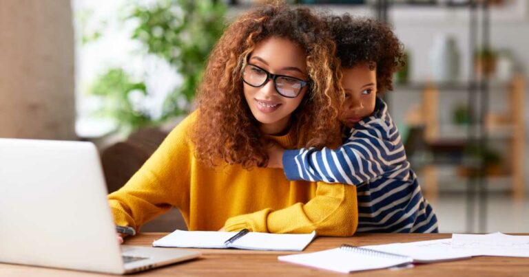 Mulher usando óculos e suéter amarelo trabalha em um notebook enquanto é abraçada por uma criança pequena com blusa listrada azul e branca, aparentando ser seu filho. Ambos estão sentados à mesa com cadernos e papéis ao redor, em um ambiente doméstico iluminado e aconchegante, sugerindo a conciliação entre trabalho remoto e maternidade. (filhos)
