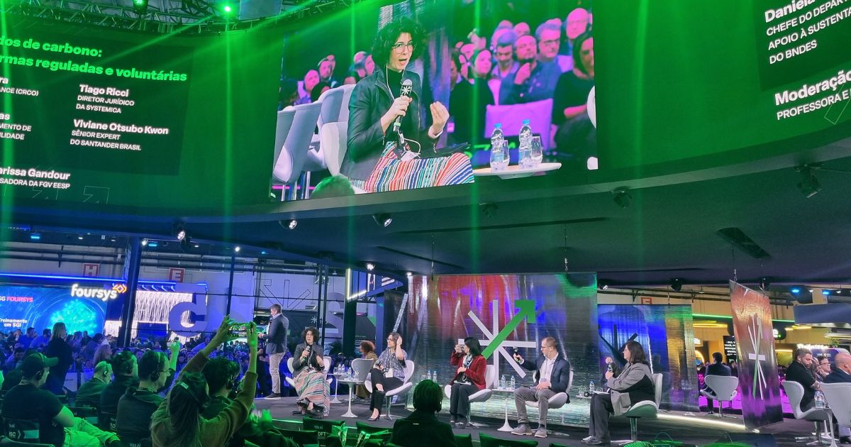 Painel de debate durante a Febraban Tech, com seis pessoas sentadas no palco discutindo o mercado de carbono. Em destaque no telão está uma das painelistas falando ao microfone, usando uma saia colorida e blazer preto. No canto superior esquerdo da tela, aparecem os nomes dos participantes: Tiago Ricci, Viviane Otsubo Kwon, Daniel Bocayuva e Larissa Gandour, além da moderação feita por professora da FGV. A plateia está cheia e atenta, em um ambiente moderno e iluminado com luzes verdes e violetas. Elementos visuais do evento, como logotipos e telas digitais, compõem o fundo da cena.