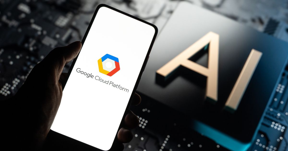 Uma mão segura um smartphone que exibe o logotipo e o texto da Google Cloud Platform. Ao fundo, vê-se a palavra "AI" (inteligência artificial) em destaque sobre uma superfície que lembra um circuito eletrônico ou uma placa tecnológica, sugerindo um ambiente digital e de alta tecnologia.
