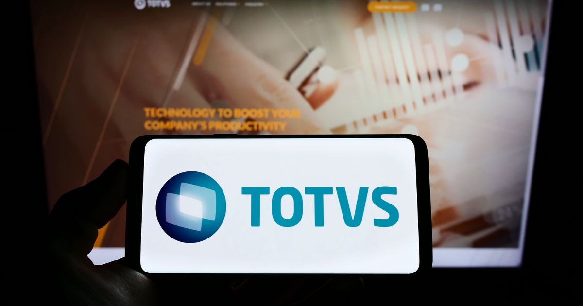 Um smartphone exibindo o logotipo da TOTVS, composto por um design geométrico azul e branco ao lado do texto 'TOTVS' em azul. Ao fundo, há uma tela de computador desfocada com o texto 'TECHNOLOGY TO BOOST YOUR COMPANY'S PRODUCTIVITY.