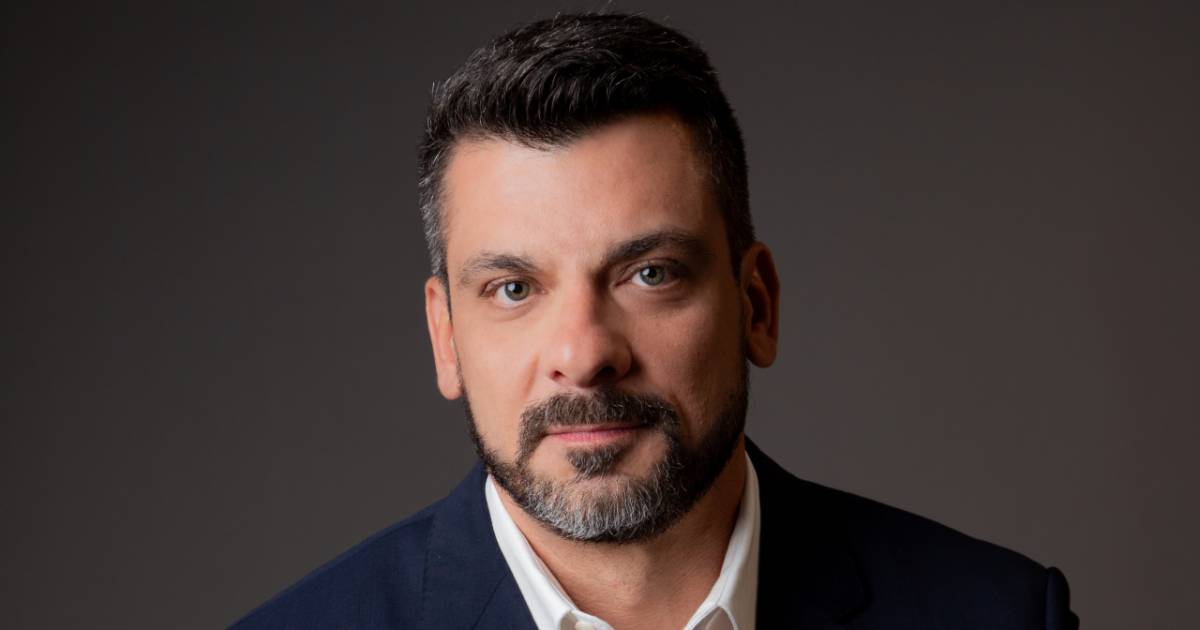 Homem de cabelo e barba curtos e escuros, usando paletó azul-marinho e camisa branca, posando em frente a um fundo neutro. Na imagem está Renato Simões, diretor de Produtos e Marketing da Oi Soluções.