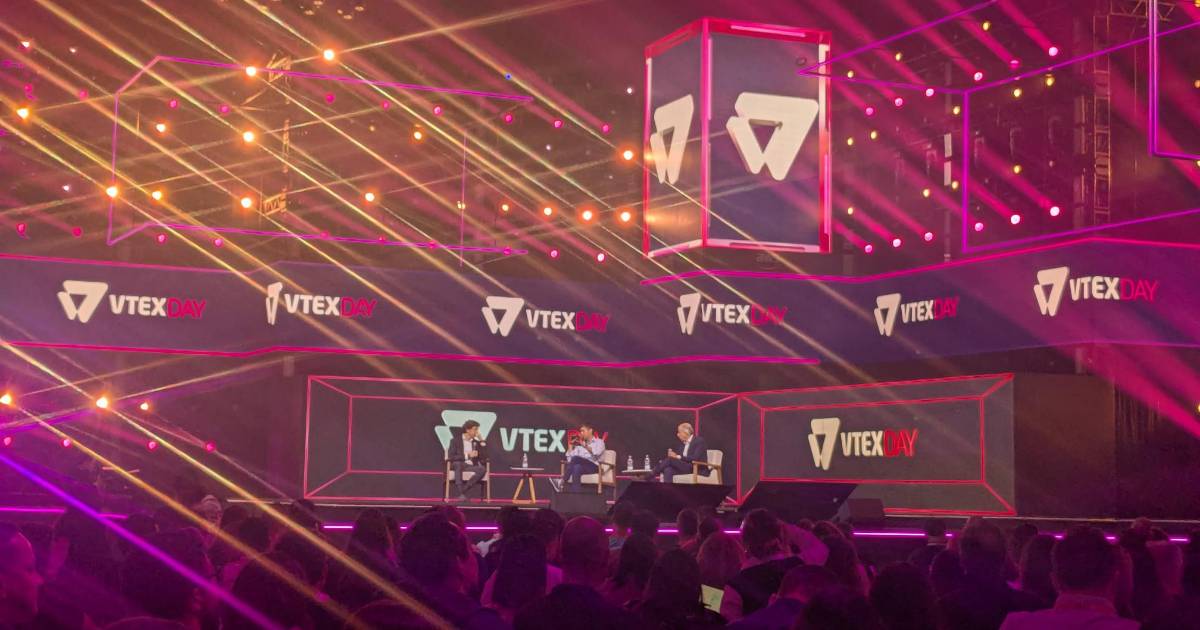 Imagem do VTEX Day com três pessoas sentadas no palco principal, participando de um painel. O cenário é iluminado com luzes coloridas e decorado com painéis digitais exibindo o logotipo do evento. Na imagem estão Alberto Serrentino, fundador da Varese Retail, Fred Trajano, CEO da Magazine Luiza, e Marcilio D'Amico Pousada, Presidente do Conselho de Administração da RD Saúde. O público está sentado em frente ao palco, acompanhando a discussão. (Magalu)