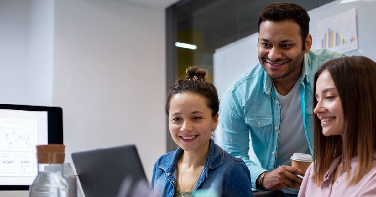 Descrição da imagem para campo "Texto alternativo" do WordPress: Três jovens profissionais sorrindo e olhando para a tela de um laptop em um ambiente de escritório moderno. Ao fundo, há uma parede de vidro e um quadro branco com gráficos impressos. Um homem de camisa azul clara segura um copo de café enquanto interage com duas mulheres, uma de camisa jeans e outra de blusa rosa claro. Um monitor com gráficos de dados também é parcialmente visível à esquerda da imagem. Contexto: Porto Digital.