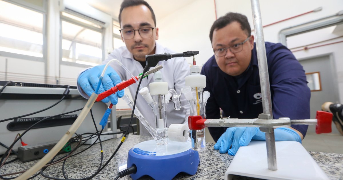 Imagem mostra dois pesquisadores em um laboratório da Unicamp manuseando equipamentos científicos. Ambos usam jalecos, luvas azuis e óculos de proteção. Um dos pesquisadores ajusta conexões elétricas em um aparato de vidro conectado a um agitador magnético azul sobre uma bancada. O ambiente é bem iluminado, com janelas amplas ao fundo e equipamentos laboratoriais visíveis, caracterizando uma atividade de pesquisa científica.