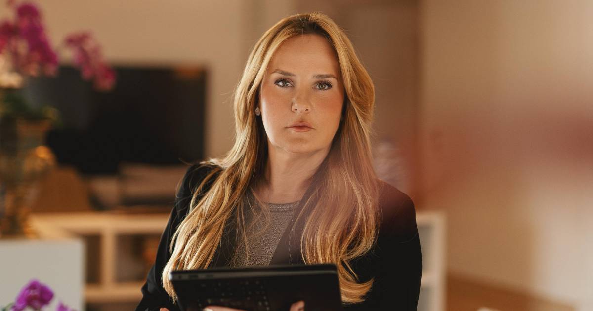 Mulher loira de cabelos longos e lisos, vestindo roupa social escura, segura um tablet e olha diretamente para a câmera. Ao fundo, há um ambiente interno com móveis e flores decorativas. Na imagem está Flávia Pollo Nassif, CEO da DMA