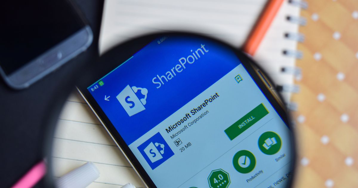 Um smartphone exibe a página de instalação do aplicativo Microsoft SharePoint na Google Play Store. Uma lupa amplia a tela, destacando informações como o nome do app, o ícone, o desenvolvedor (Microsoft Corporation), a classificação etária (3+), o tamanho do arquivo (20 MB) e os botões de instalação e avaliação. Ao fundo, há um caderno com espiral laranja e uma caneta branca, todos sobre uma superfície preta.
