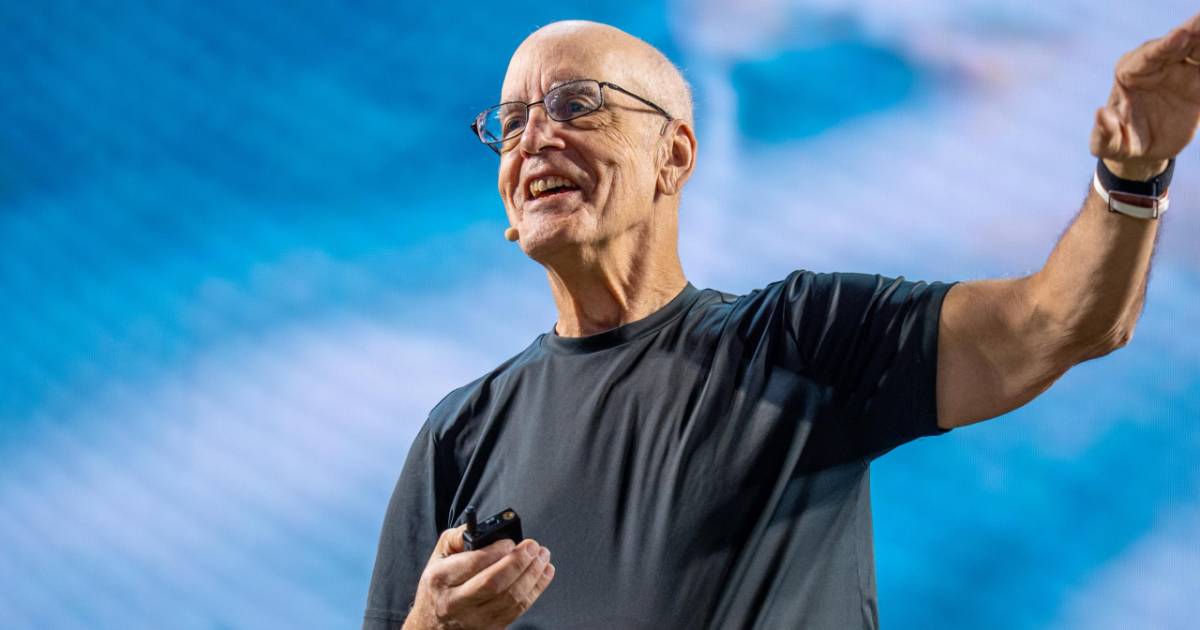 Homem careca e de óculos, sorrindo e falando em um palco durante palestra, usando camiseta preta e microfone de lapela. Ele segura um controle remoto pequeno na mão direita e gesticula com o braço esquerdo levantado. Ao fundo, há um painel desfocado em tons de azul. Trata-se de Gary Bolles, especialista em inteligência artificial, no IT Forum Praia do Forte 2025. (instituto)