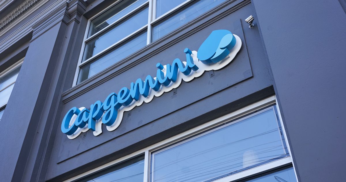 A imagem mostra a parte externa de um edifício com uma placa que exibe o nome "Capgemini" em letras cursivas azuis, acompanhadas de um logotipo azul e branco. A placa está fixada na fachada do prédio, que possui grandes janelas. (tendencias)
