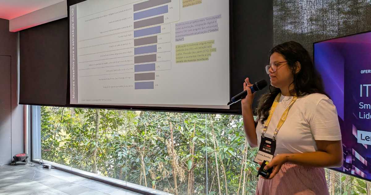 Bruna Bomfim, gerente de Inteligência da Itaqui, apresentando palestra durante o IT Forum Na Mata, em frente a um telão com gráficos e segurando um microfone. (investimentos)