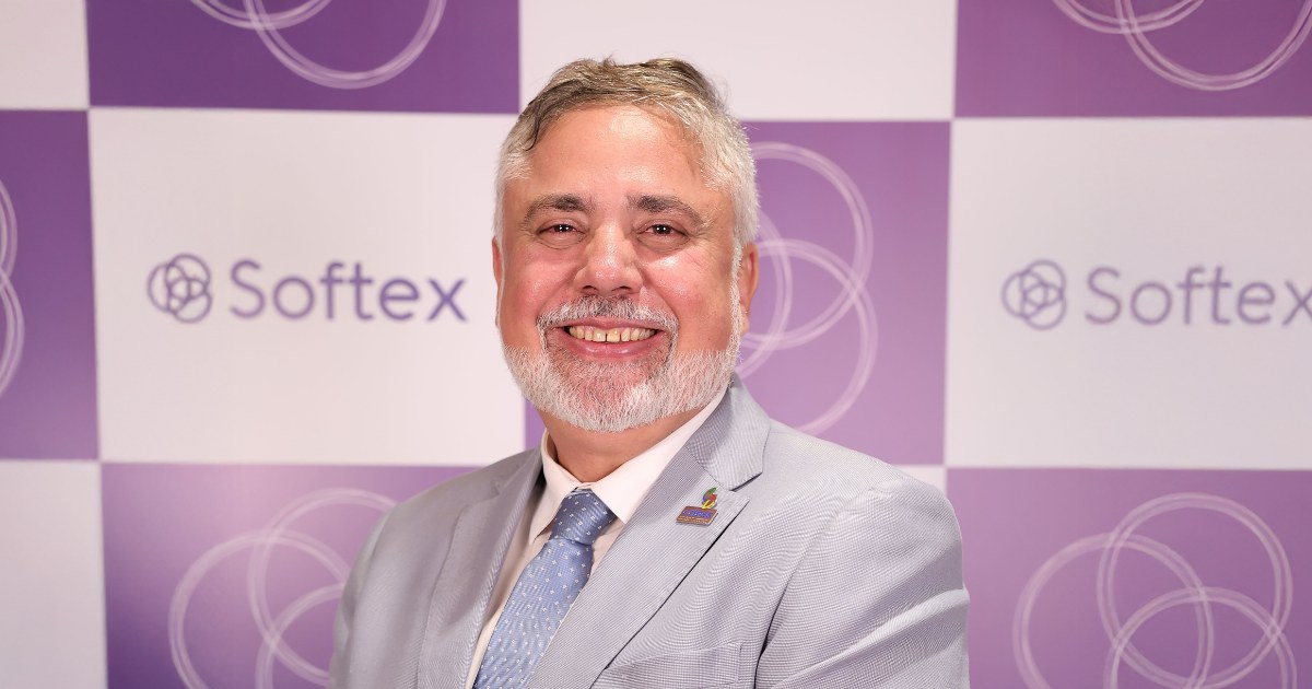 Esta imagem mostra uma pessoa sorridente, vestindo um terno claro com gravata azul. O fundo tem um padrão quadriculado com quadrados roxos e brancos alternados, cada um contendo o logotipo e o nome da Softex — o que sugere que a foto foi tirada em um evento ou ambiente institucional ligado à organização.