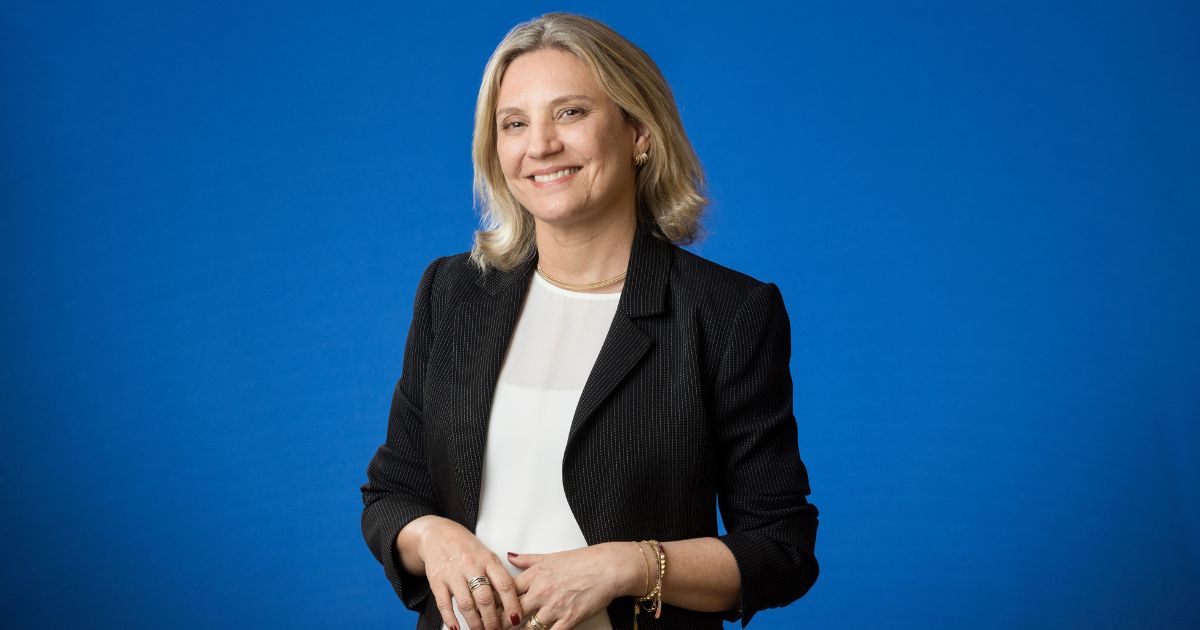 Pessoa em pé diante de um fundo azul sólido, vestindo blazer preto sobre uma blusa branca. Tem cabelo claro, está sorrindo e com as mãos entrelaçadas à frente do corpo. (tecnologia)