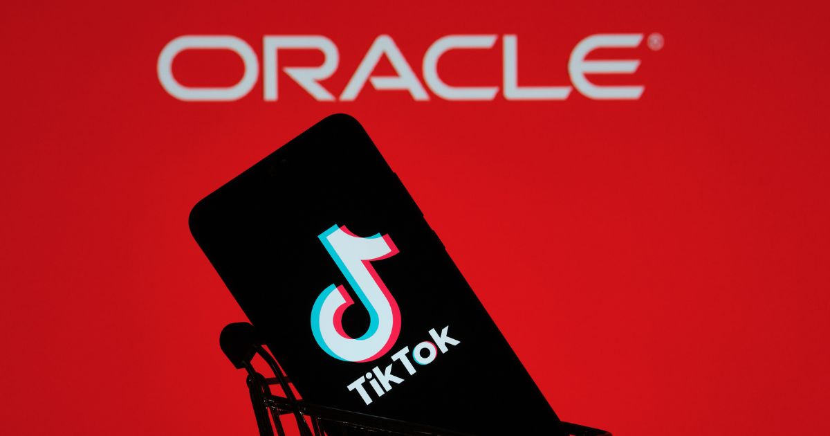 Um smartphone exibindo o logotipo e o nome do TikTok está posicionado dentro de um pequeno carrinho de compras. Ao fundo, vê-se um fundo vermelho com a palavra "ORACLE" escrita em letras brancas, sugerindo uma relação comercial ou tecnológica entre as marcas.
