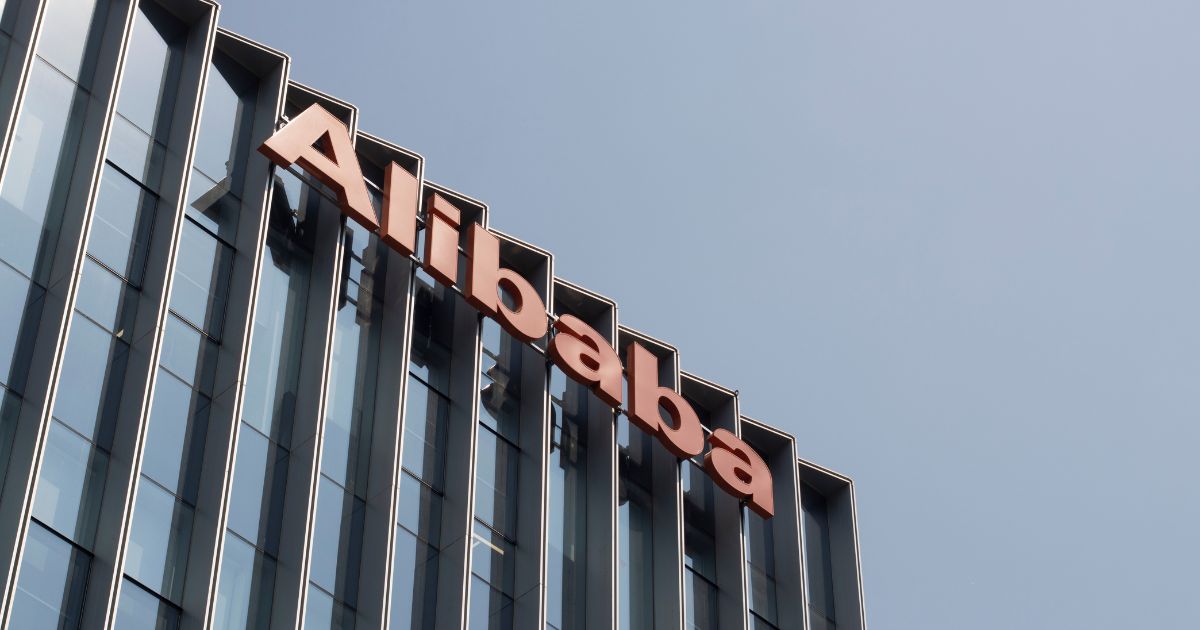 Fachada de um edifício contemporâneo com design limpo e janelas verticais alinhadas. No topo da estrutura, destaca-se uma placa com a inscrição "Alibaba" em letras grandes e na cor laranja vibrante. O céu ao fundo está claro, sem nuvens, contribuindo para uma atmosfera iluminada e profissional. A imagem transmite a presença marcante da marca Alibaba, sugerindo tratar-se de uma sede ou unidade corporativa da empresa.