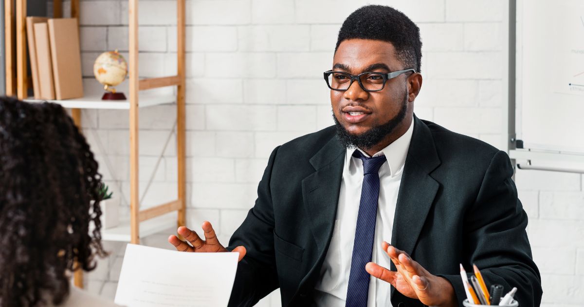 Um homem vestido com terno e gravata, usando óculos e com cabelo e barba bem aparados, está em um ambiente profissional, aparentemente envolvido em uma conversa com outra pessoa que está de costas para a câmera. Ao fundo, há prateleiras com livros, um globo terrestre e parte de uma lousa branca visível à direita. A cena sugere uma reunião de negócios ou uma entrevista em um escritório bem organizado. (emprego/vagas)