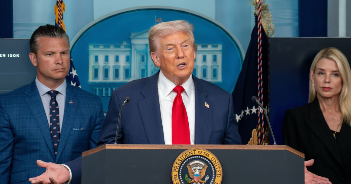 Três pessoas estão posicionadas atrás de um púlpito que exibe o selo do Presidente dos Estados Unidos. A pessoa ao centro está falando ao microfone, vestindo um terno escuro, camisa branca e gravata vermelha. Ao fundo, há uma imagem da Casa Branca e duas bandeiras. À esquerda, uma pessoa veste um terno azul xadrez com camisa branca e gravata estampada. À direita, outra pessoa está com uma roupa preta. (Trump e Microsoft)