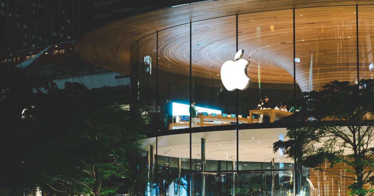 A imagem mostra uma loja da Apple com arquitetura marcante. A fachada é feita de vidro curvado e o teto interno tem um padrão circular de madeira. O logotipo da Apple está iluminado e centralizado na frente da loja. Dentro, há mesas exibindo produtos da marca e várias pessoas interagindo com os dispositivos ou entre si. A loja está cercada por árvores e parece estar localizada em uma área urbana à noite, como indicado pela iluminação e pelos reflexos no vidro. (Apple)
