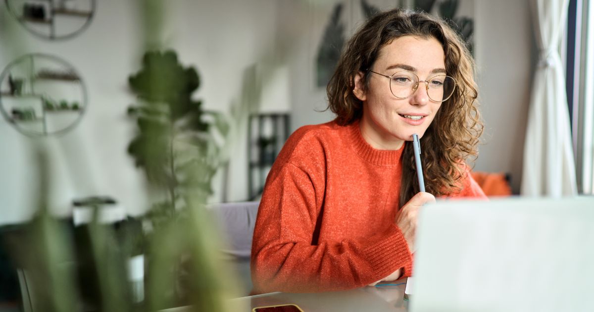 Uma pessoa com cabelo cacheado e óculos está sentada à mesa, vestindo um suéter laranja. Ela segura uma caneta próxima à boca enquanto observa atentamente a tela de um laptop à sua frente. Ao fundo, é possível ver um ambiente interno desfocado, com plantas e prateleiras na parede, sugerindo um espaço de trabalho ou estudo. A imagem transmite concentração e envolvimento, possivelmente durante uma reunião online ou momento de estudo de cursos.