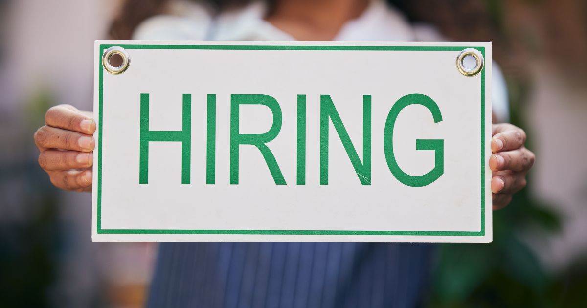A imagem mostra uma pessoa segurando uma placa retangular branca com bordas verdes e dois ilhós metálicos nos cantos superiores. No centro da placa, em letras grandes e verdes, está escrito “HIRING”. A pessoa está usando uma roupa com listras verticais azuis e brancas, e o fundo é desfocado, sugerindo um ambiente externo com vegetação. (oportunidades, vagas)