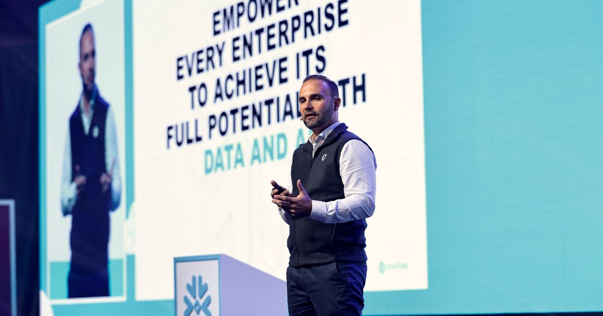 A imagem mostra uma pessoa em pé sobre um palco, vestindo camisa branca e colete preto com um logotipo visível no lado esquerdo do peito. A pessoa está segurando um dispositivo pequeno nas mãos, sugerindo que está fazendo uma apresentação. Ao fundo, há um grande painel com texto em inglês que diz: “EMPOWER EVERY ENTERPRISE TO ACHIEVE ITS FULL POTENTIAL WITH DATA AND AI” O cenário é iluminado com tons claros e azulados, típico de eventos corporativos ou conferências de tecnologia. À esquerda, há uma tela adicional exibindo a mesma pessoa, reforçando que se trata de uma apresentação em grande escala. (Snowflake)