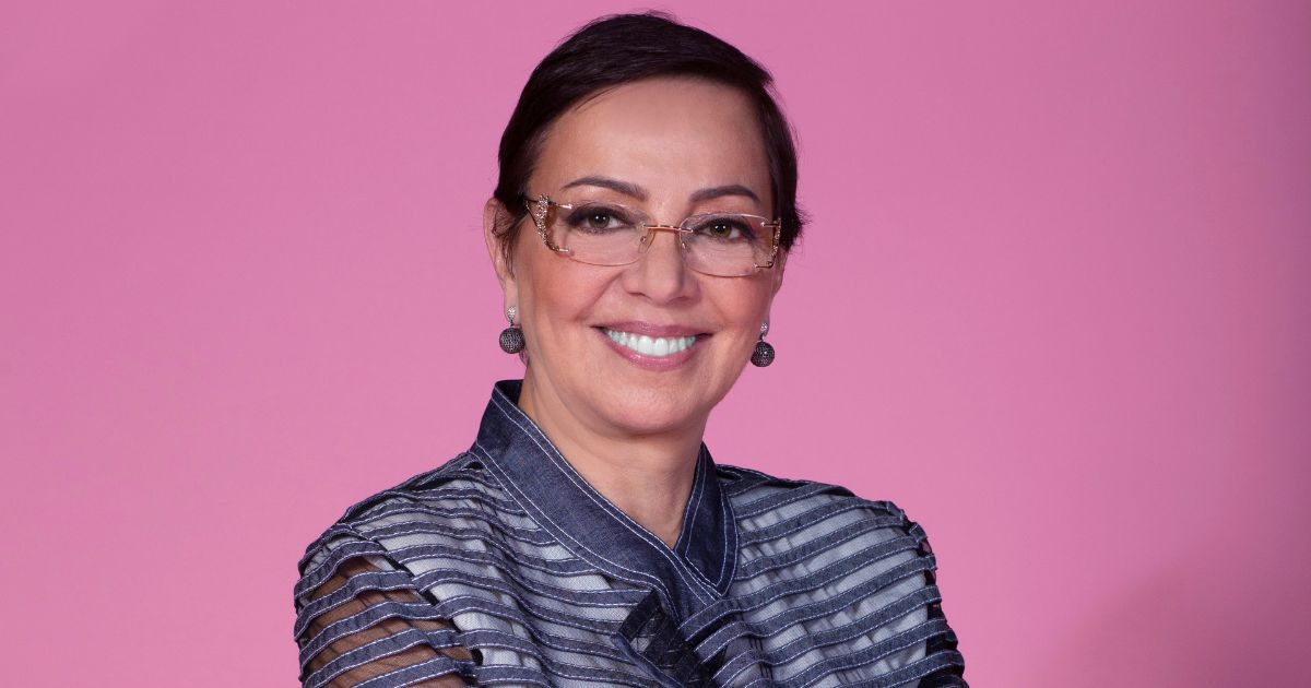 A imagem mostra uma pessoa vestindo uma jaqueta estilosa de tecido semelhante ao jeans, com um design moderno composto por faixas horizontais translúcidas nas mangas e no corpo. A peça tem gola alta estruturada e detalhes que criam um visual sofisticado e contemporâneo. O fundo é totalmente rosa, liso e uniforme, destacando ainda mais a roupa e criando um contraste vibrante. A pessoa está posicionada de frente, com os braços cruzados, transmitindo uma postura firme e elegante. (Tânia)