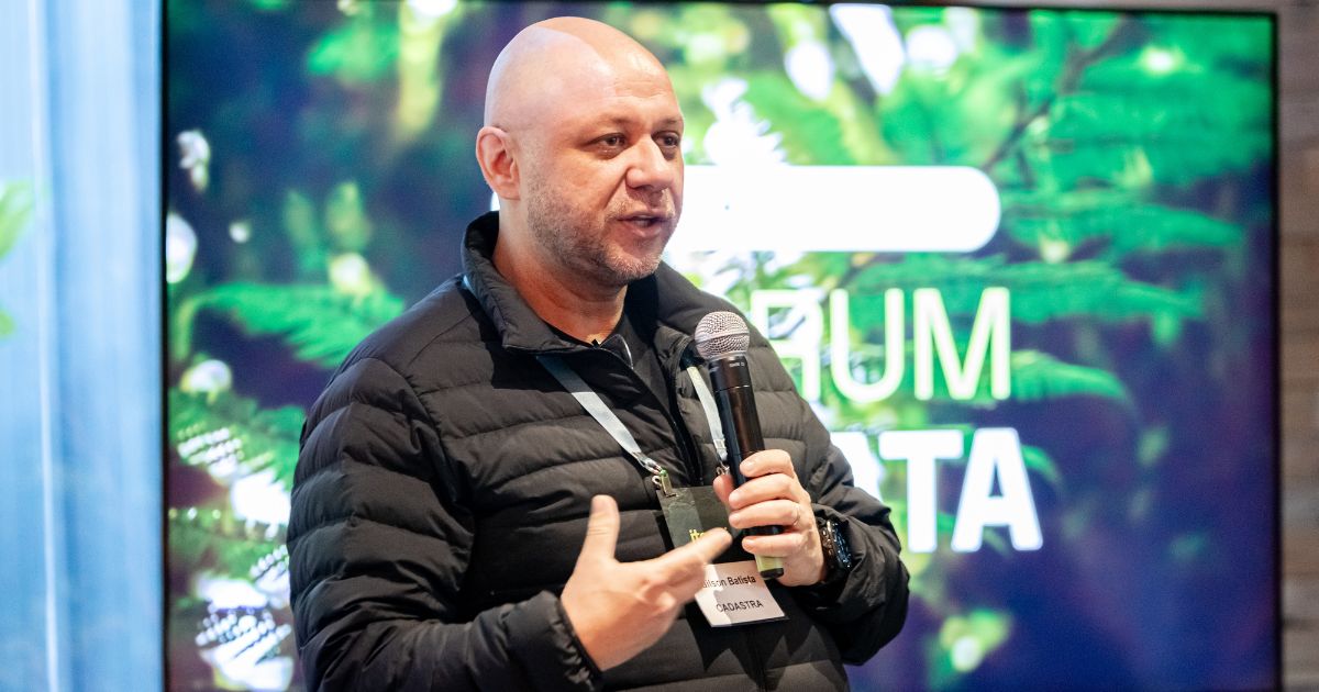 A imagem mostra uma pessoa vestindo um casaco acolchoado preto e um crachá pendurado no pescoço, segurando um microfone com uma das mãos enquanto faz um gesto com a outra. Ao fundo, há uma tela grande exibindo uma imagem de folhas verdes, sugerindo um tema relacionado à natureza. Sobre a tela, é possível ler parcialmente um texto em letras grandes, onde se destacam as palavras “FÓRUM” e “MATA”. A iluminação é clara e o ambiente parece ser um espaço interno voltado para apresentações ou palestras. (IA)