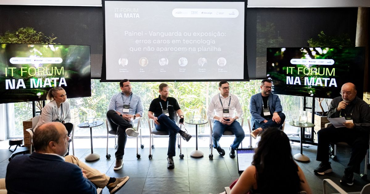 A cena mostra um painel de discussão em um evento chamado IT Forum na Mata. Há seis pessoas sentadas em cadeiras dispostas em linha, com uma pessoa adicional sentada mais à frente, de costas para a câmera. Cada participante usa crachá e está em um ambiente iluminado por luz natural, com grandes janelas ao fundo revelando vegetação externa. No centro da imagem, há um telão com o título do painel: “Painel – Vanguarda ou exposição: erros caros em tecnologia que não aparecem na planilha”. Abaixo do título, aparecem ícones e nomes dos palestrantes. À esquerda e à direita do telão, existem duas telas adicionais exibindo o logotipo do evento: “IT FORUM NA MATA” sobre um fundo verde com folhas. No chão, há piso escuro e algumas mesas pequenas com garrafas de água. A pessoa mais próxima da câmera parece estar tomando notas ou acompanhando a discussão com um dispositivo eletrônico.