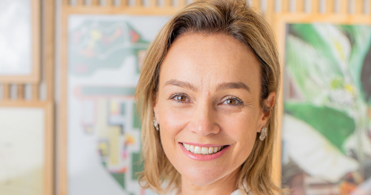 A imagem mostra um retrato em close de uma pessoa com cabelo claro, sorrindo. Ela usa brincos e está posicionada diante de um fundo desfocado que contém obras de arte ou fotografias, sem destaque. A composição foca nos traços faciais da pessoa, sendo adequada para uso em contextos profissionais ou ilustrativos. (Flávia Picolo)