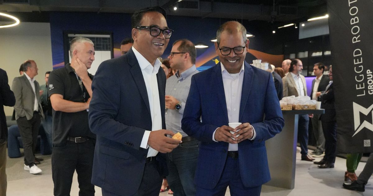 A imagem mostra um grupo de pessoas em um ambiente profissional, possivelmente durante um evento de networking ou conferência. No primeiro plano, duas pessoas vestindo ternos estão conversando e segurando bebidas. Ao fundo, outros participantes também interagem e consomem petiscos. À direita, há um banner com o texto "LEGGED ROBOT GROUP", sugerindo que o evento esteja relacionado à área de robótica ou tecnologia. O ambiente é interno, com iluminação moderna e decoração contemporânea. (TCS)